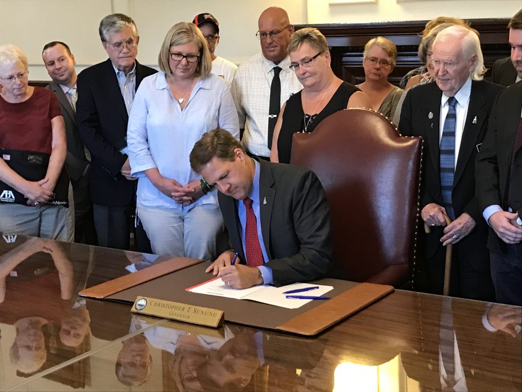 Governor Sununu signs SB282 infront of a group of on lookers and supporters.